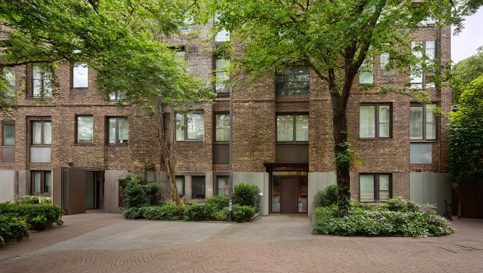 Landscaped courtyard entrance of a modern brick apartment building on a tree-lined street