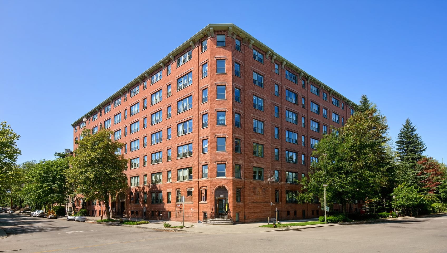 Brick corner facade of a six-storey apartment building on a tree-lined East Vancouver street under a blue sky