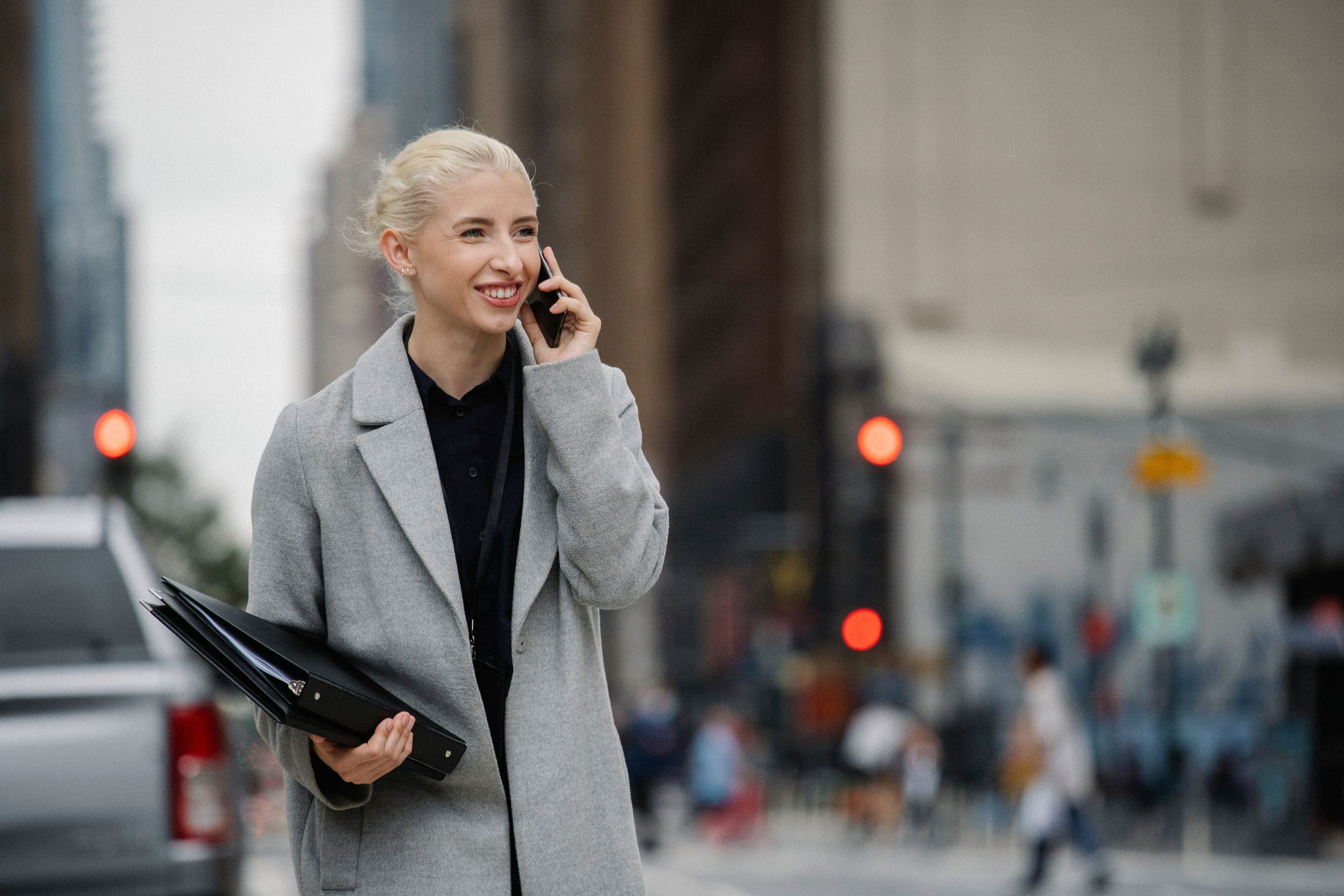 A blonde market intelligence specialist in a grey coat smiling while on a phone call on a city street