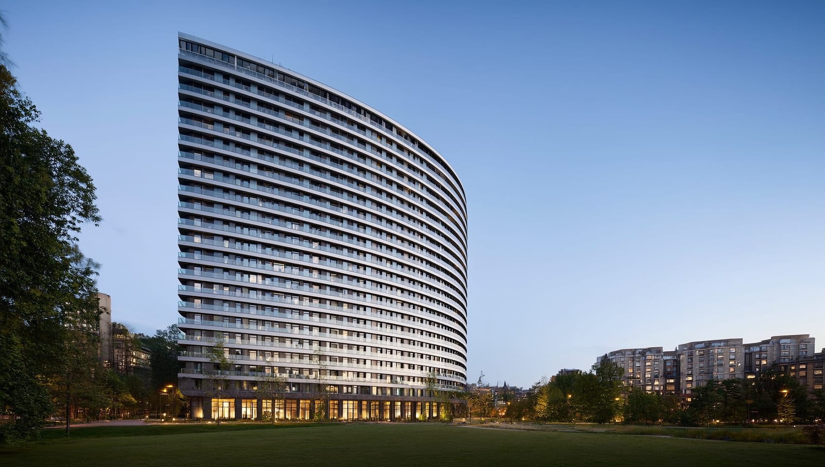 Dusk exterior photograph of a modern curved-facade high-rise residential tower beside an urban park